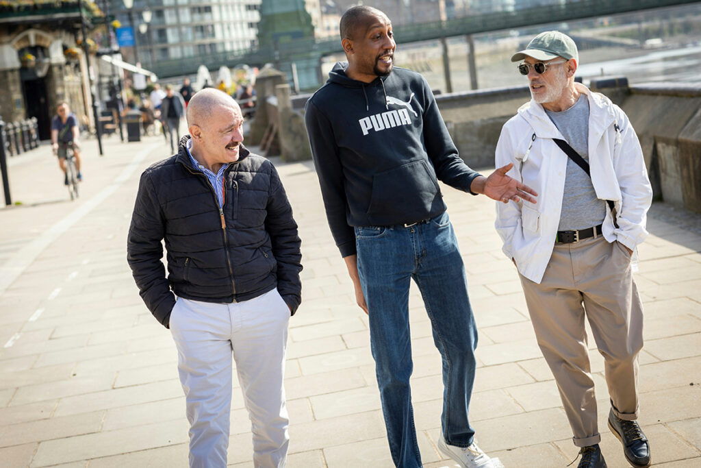 Three older men walk together on a sidewalk while having a conversation.