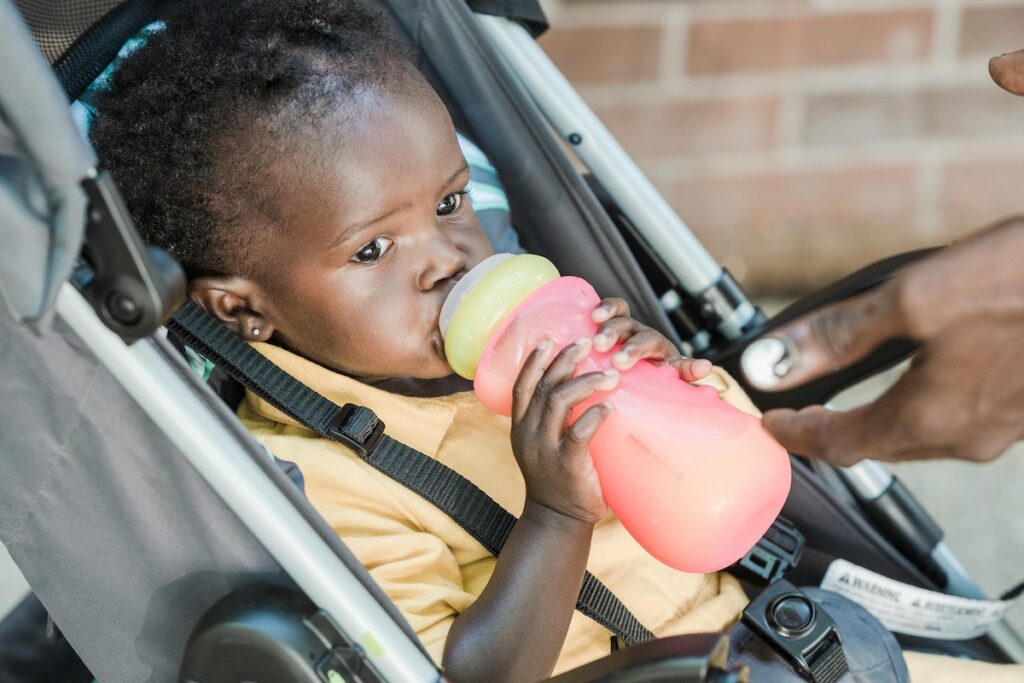A small toddler holds a pink sippy cup and licks the top of it while sitting in a stroller