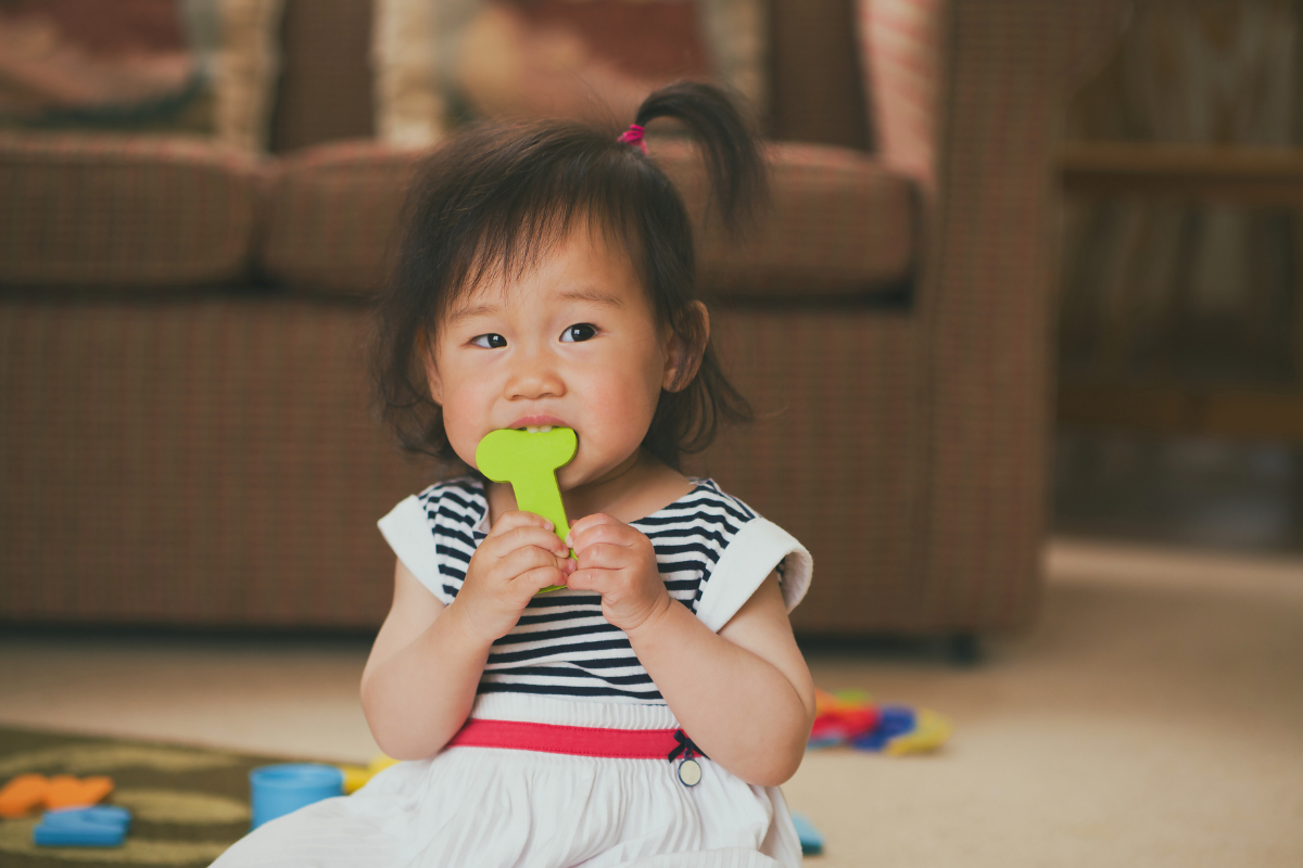 Toddler sitting down and biting a toy.