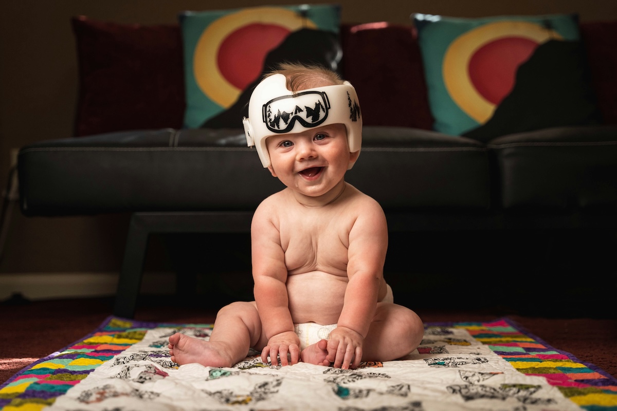 A smiling baby sitting on a floor wearing a helmet