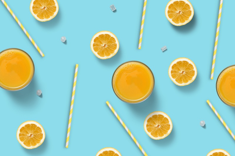 A bright blue background with an overhead arrangement of glasses of orange juice, orange straws, and orange slices.