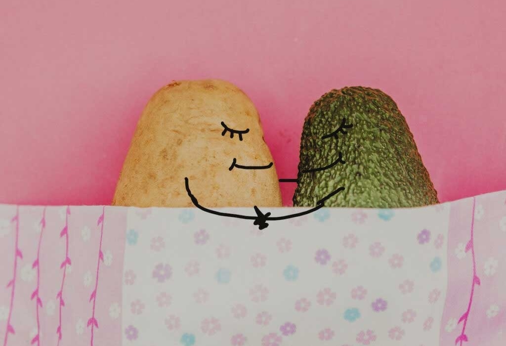 A potato and an avocado, decorated with silly faces, snuggle under are blanket.
