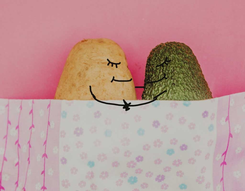 A potato and an avocado, decorated with silly faces, snuggle under are blanket.