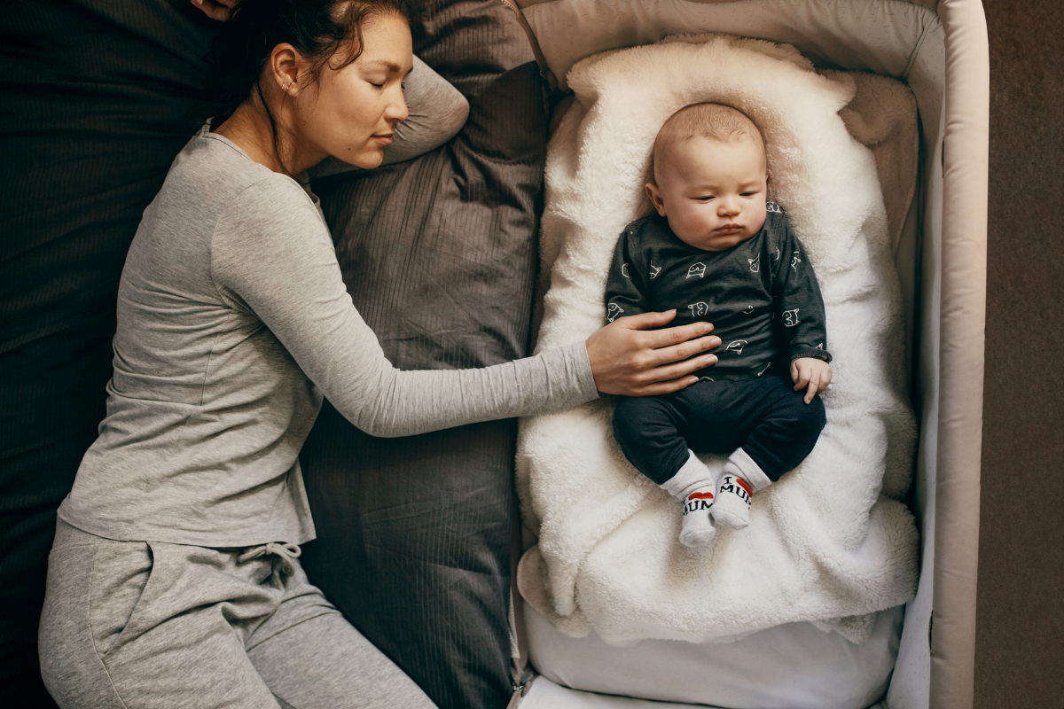 A sleeping mother with her hand on her baby's stomach as he lays next to her in his bassinet.