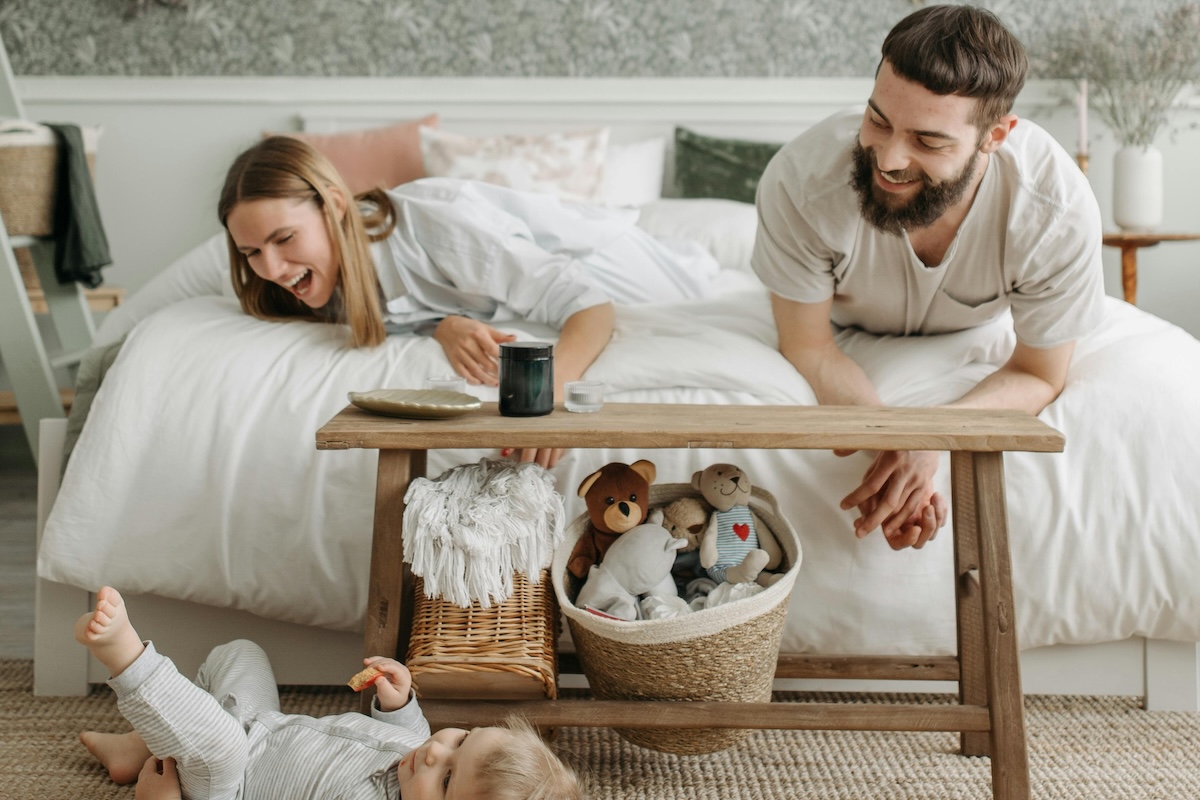 Two partners lying on bed playfully and looking at the kid on floor