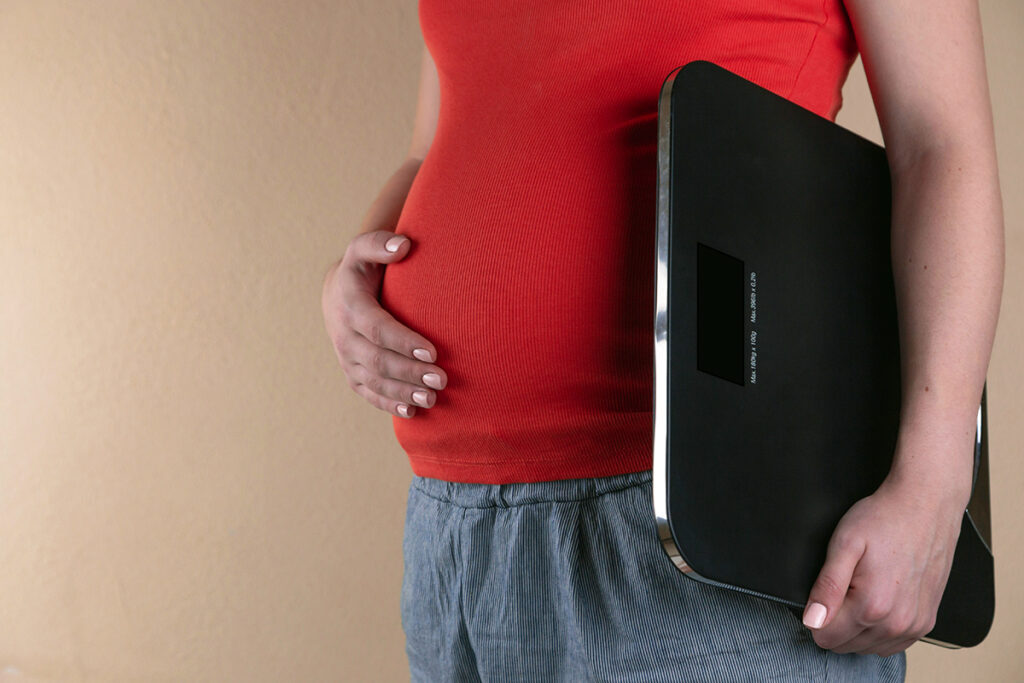 Close up of pregnant person's belly with their hand on their stomach and a scale in the other hand