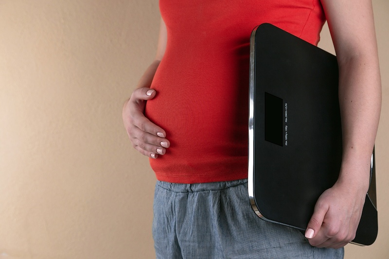 Close up of pregnant person's belly with their hand on their stomach and a scale in the other hand