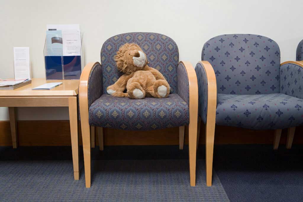 A teddy bear sits on a chair in a doctor's waiting room.