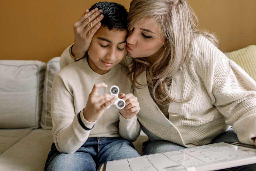 A child sits on a couch playing with a fidget spinner. His mother, seated next to him, kisses him on the forehead.