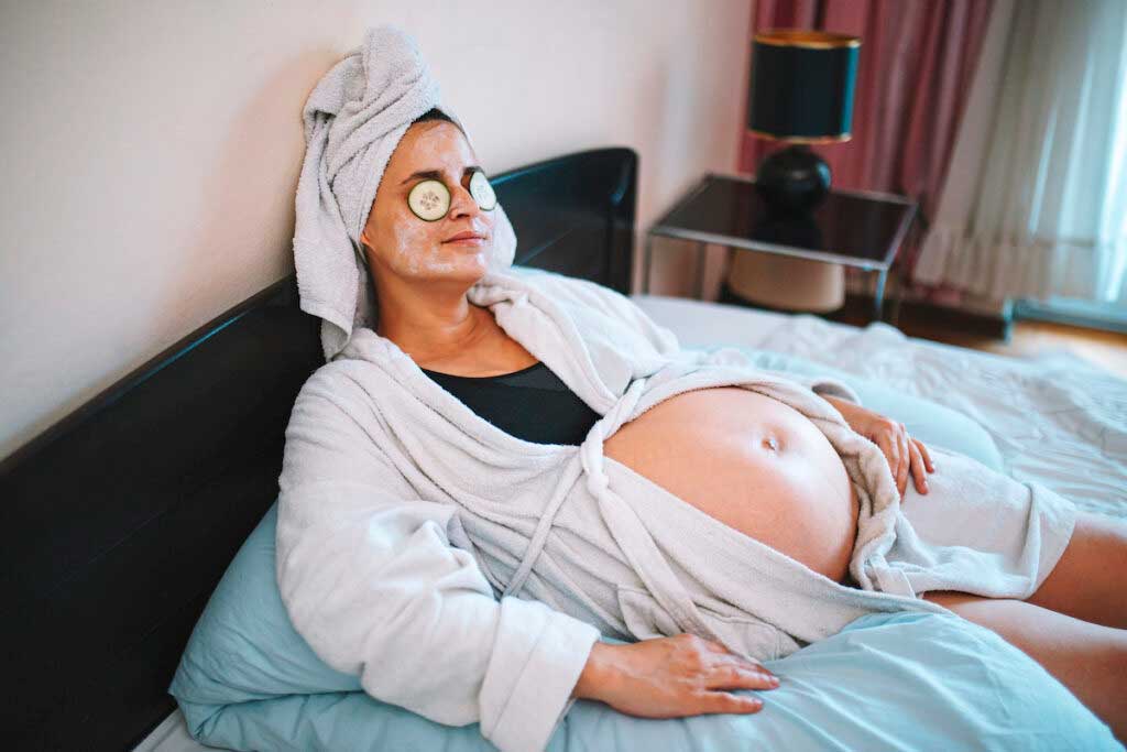 A person with a pregnant belly enjoys a spa day facial mask with cucumber eye covers.