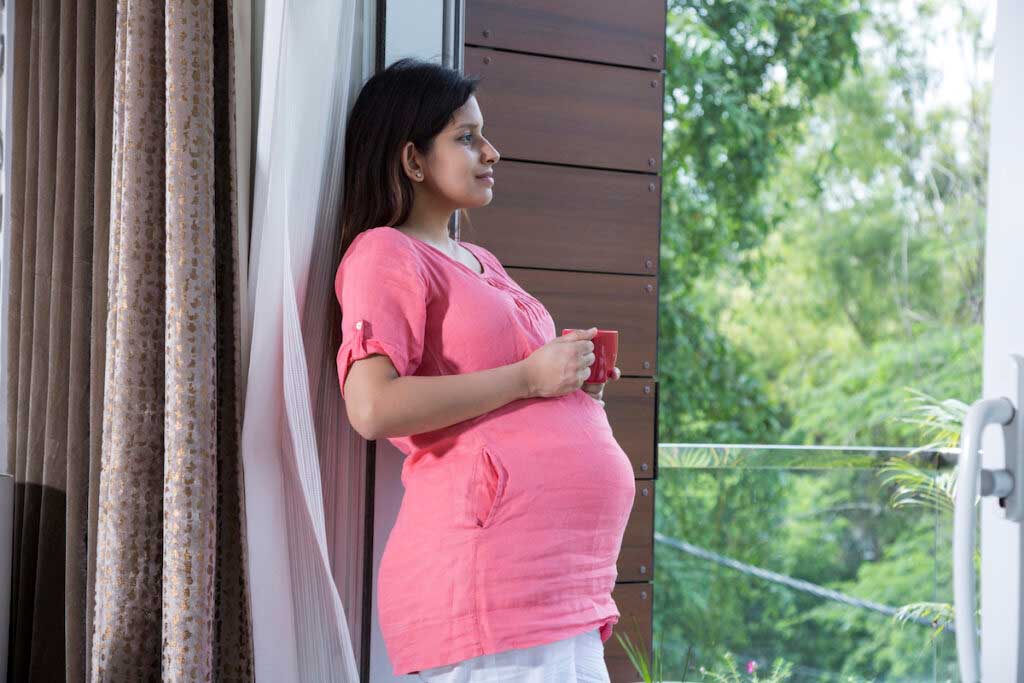 Pregnant person stands on a balcony while drinking a cup of coffee and smiling.