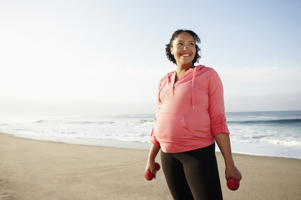 A woman in jogging pants and a hoodie smiles while standing on the beach.
