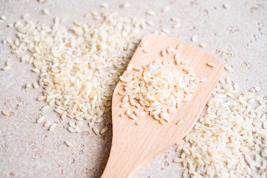Close up of uncooked rice and a wooden spoon on a tan table.