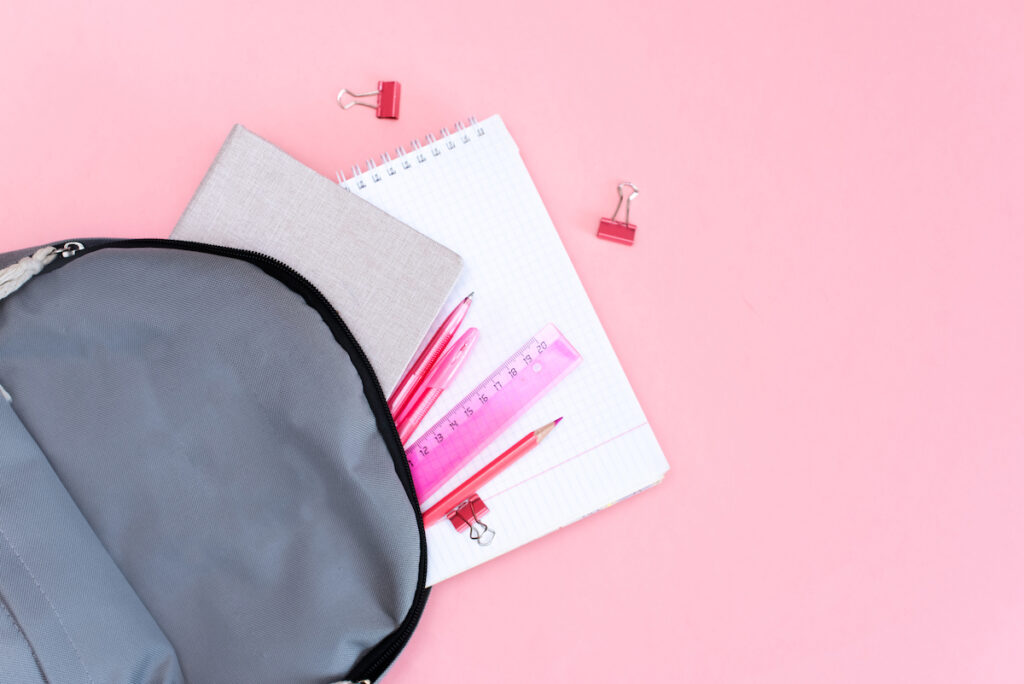 A gray backpack with contents, including paper and pink binder clips, spilled on a pink background.