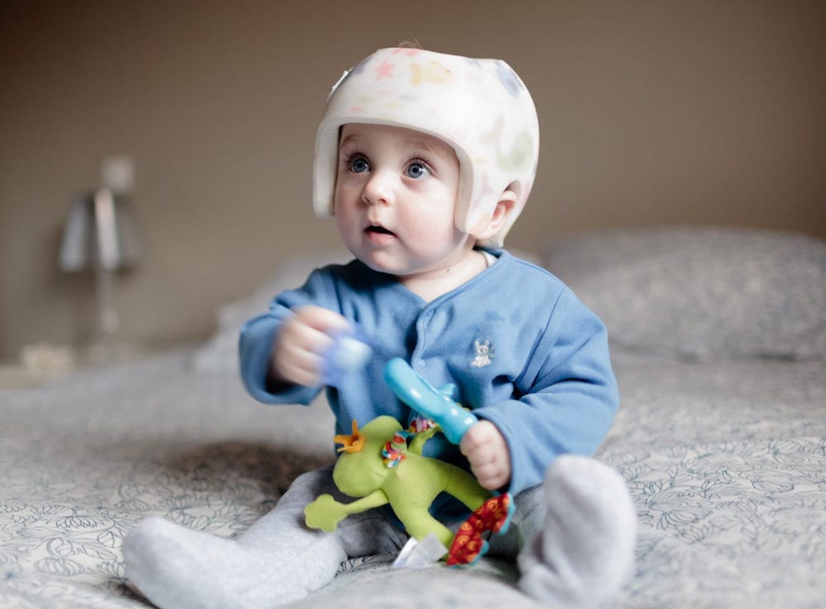 A small seated baby plays with a toy on the floor while wearing a helmet.