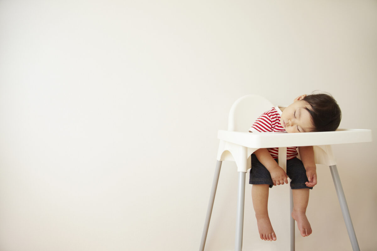 Small child sits in a highchair, sleeping. The child's head is resting on the highchair tray.