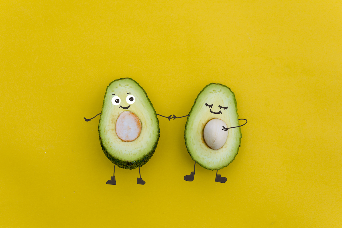 Two avocado halves, one with the seed still in, are decorated with smiling faces and arms.
