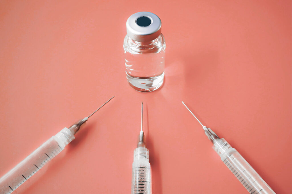Three syringes with needles point to a vial of clear liquid on a peach background.