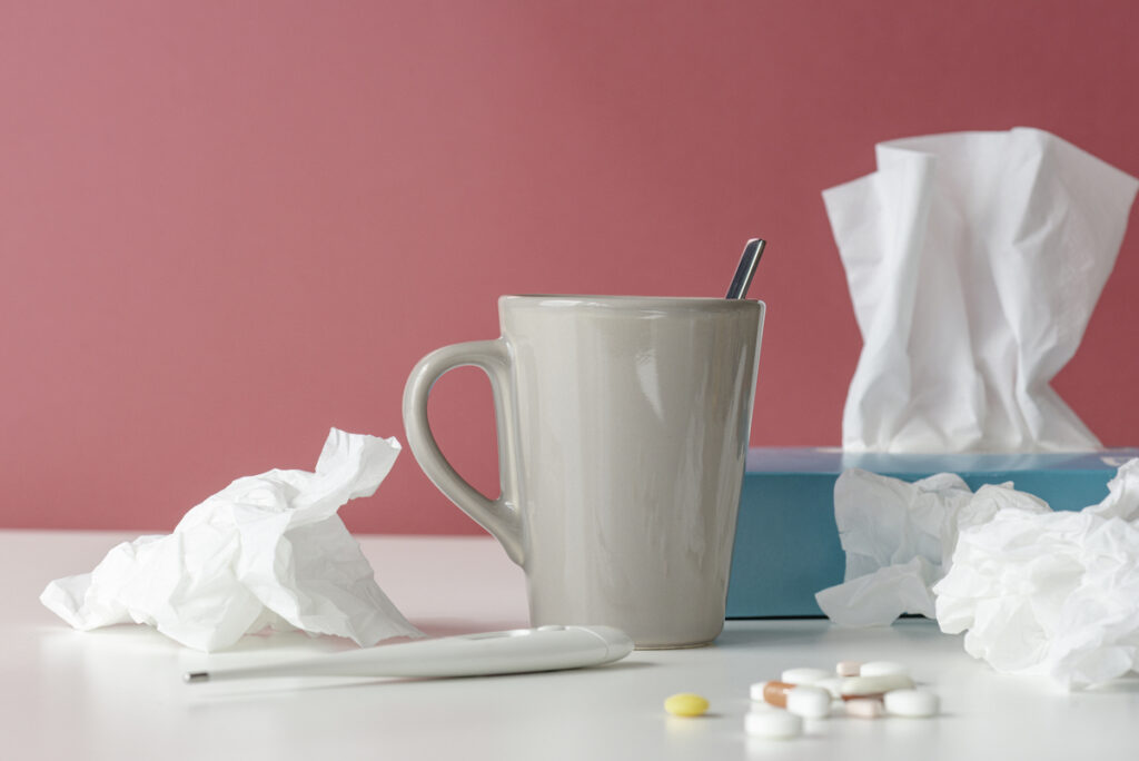 An arrangement of tissue boxes, used tissues, and a cup of hot tea against a pink background.