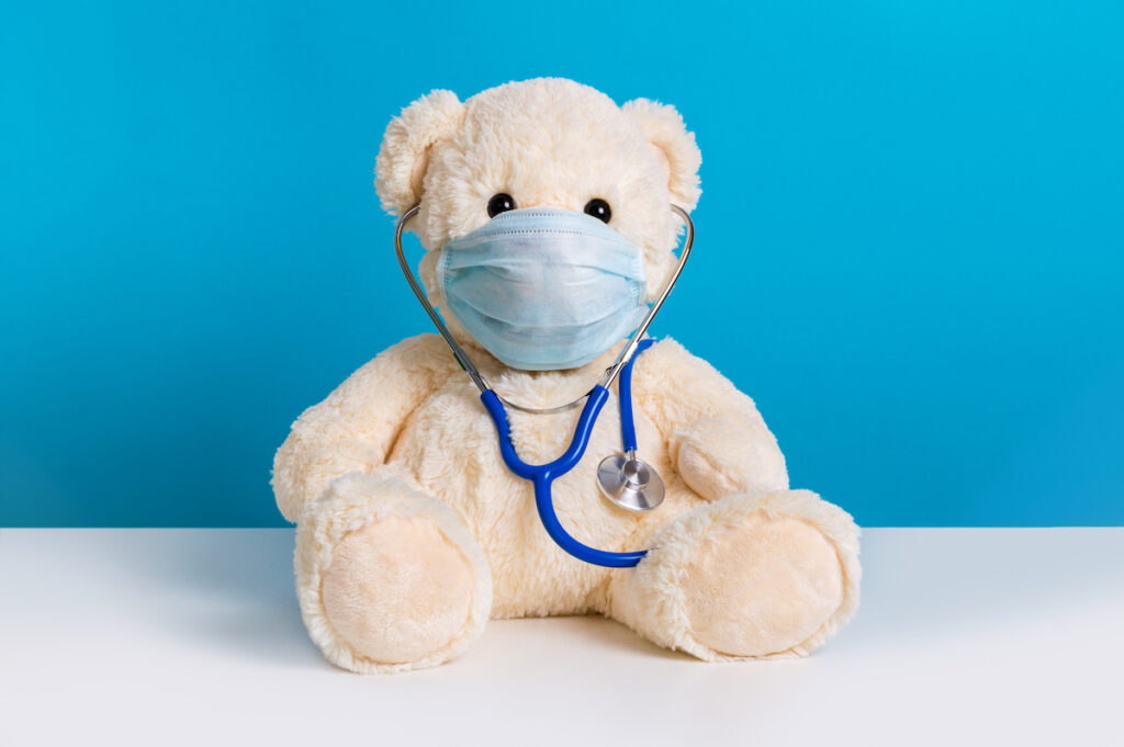 A teddy bear wears a stethoscope and mask against a blue background.