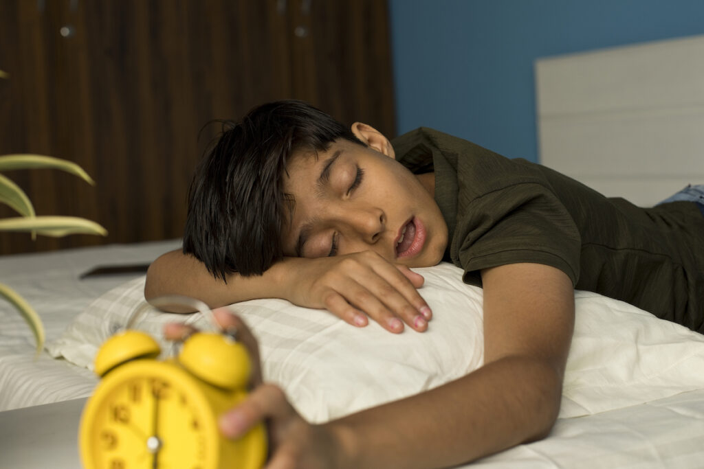 A young boy sleeping face down on a pillow reaches over to turn off a yellow alarm clock.