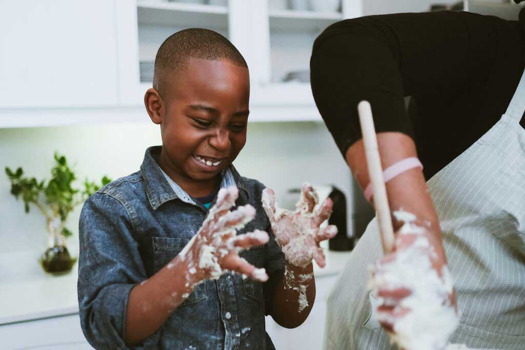 A boy with sticky dough all over his hands laughs while helping in the kitchen.