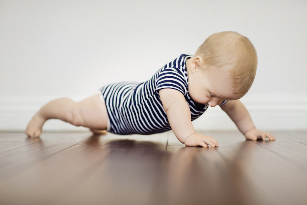 Infant in a striped onesie does a plank position on a wood floor.