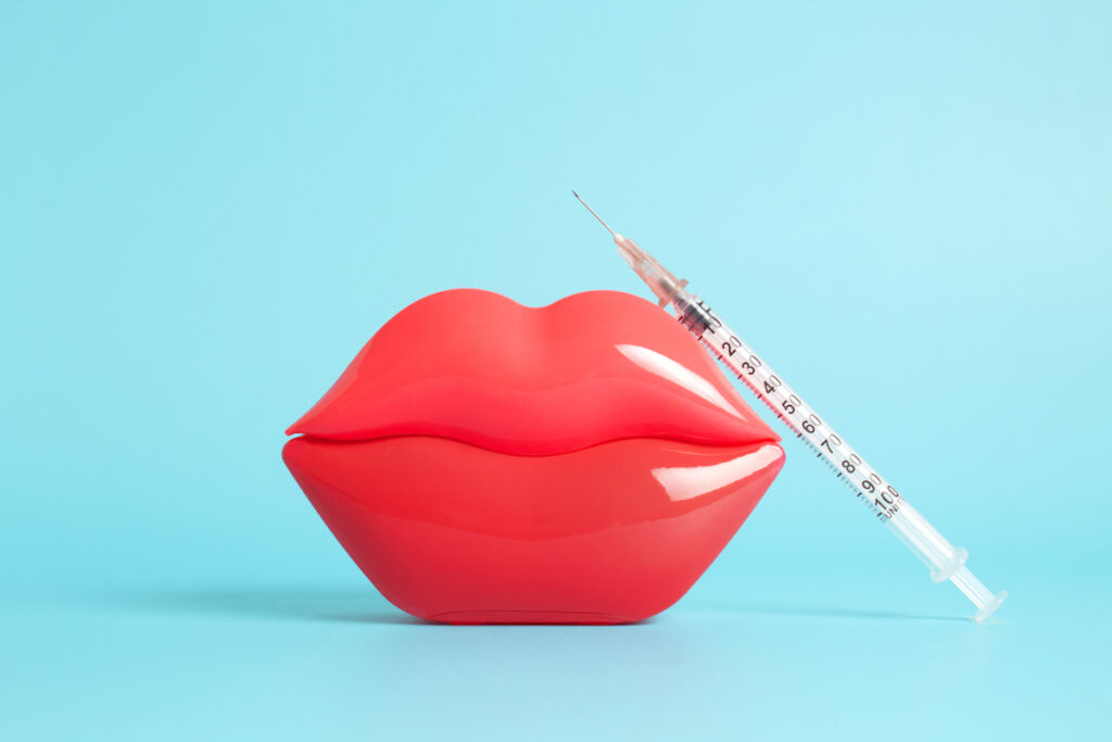 A dermal filler syringe is propped up against bright red plastic lips.