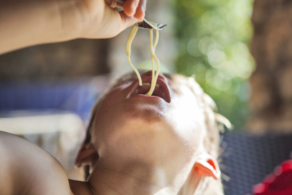 A kid tips their head back to eat a strand of spaghetti dangling from a fork.