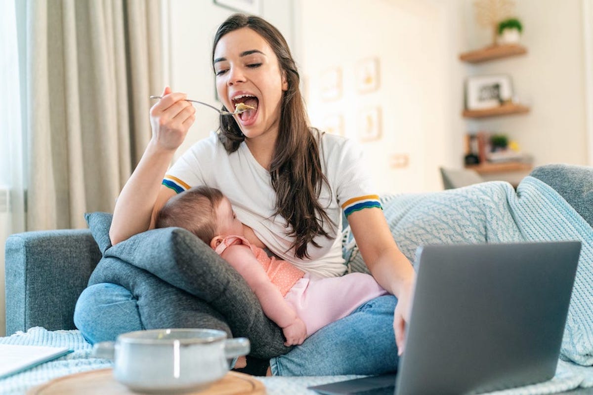 A woman breastfeeds her child while eating soup and looking at her laptop.