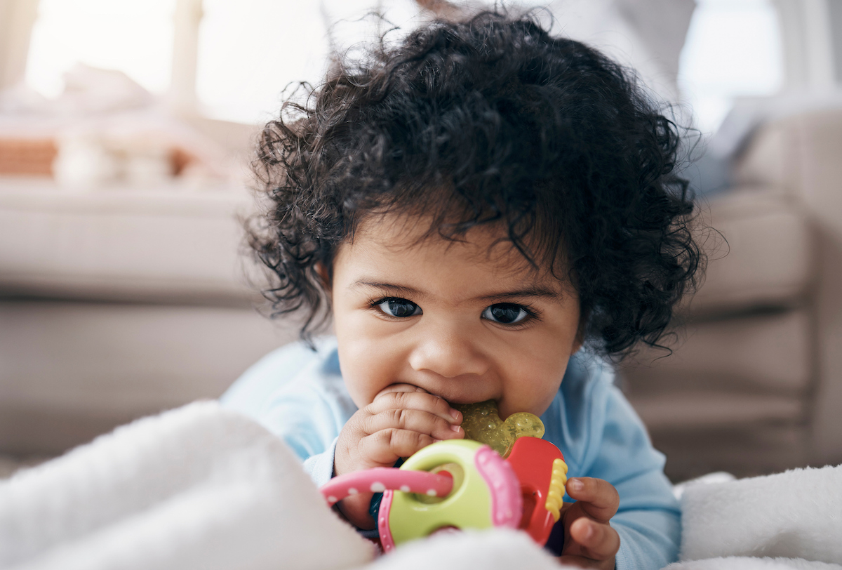 A baby lies on their tummy chewing on a teething ring and smiling.