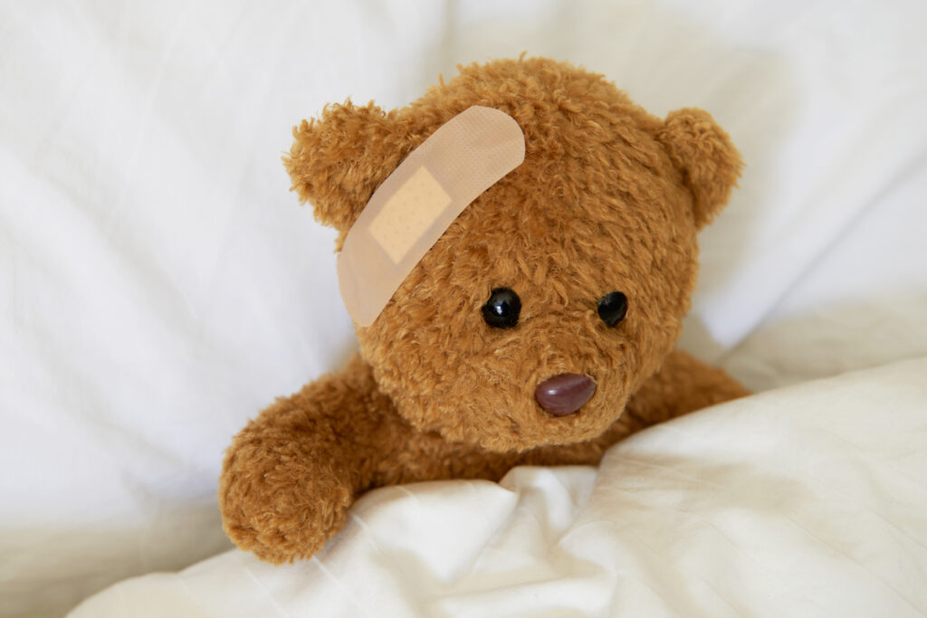 A teddy bear rests on a pillow with a bandaid across its forehead.