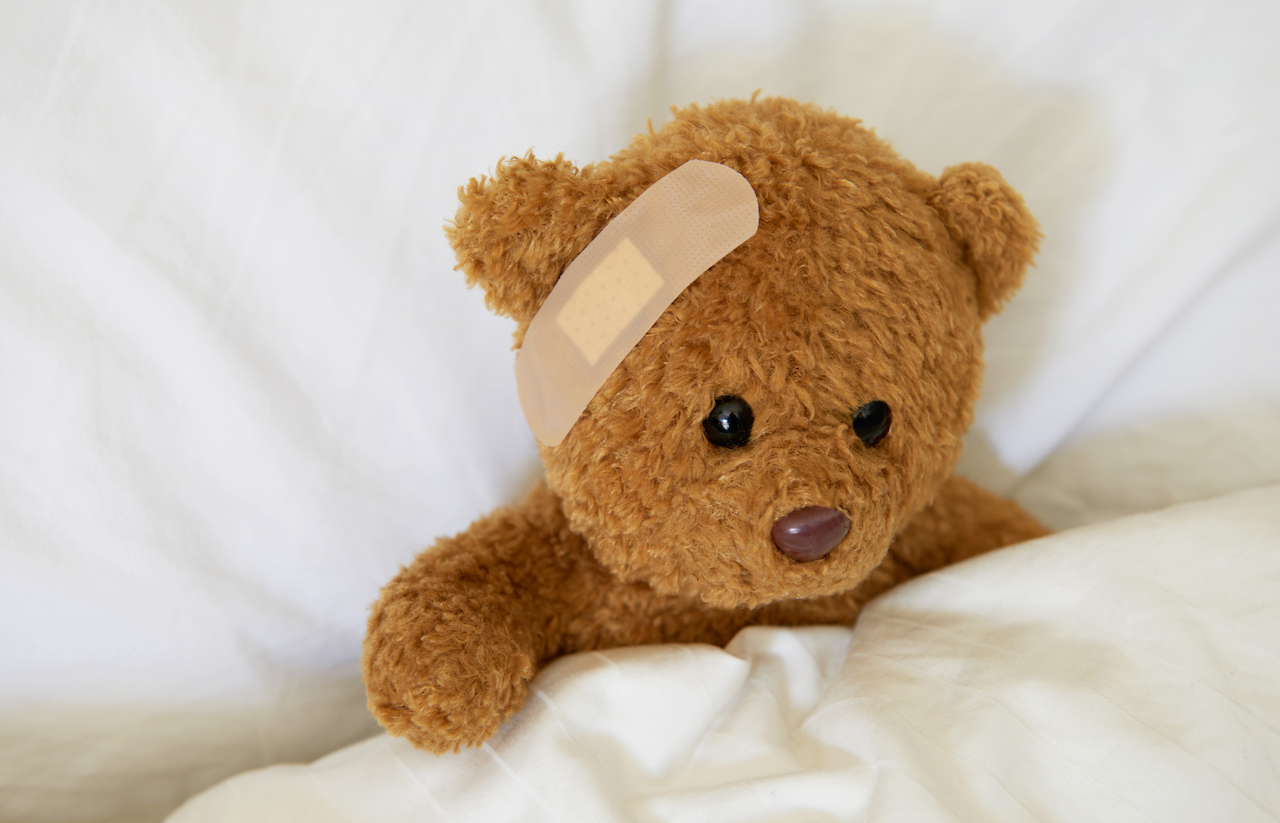 A teddy bear rests on a pillow with a bandaid across its forehead.