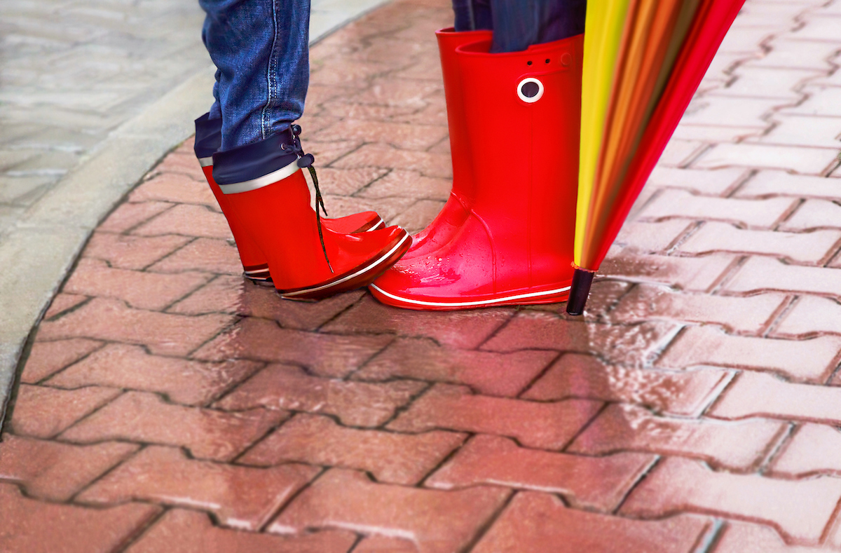 Parent and child wear red rubber boots on a rainy day. The child is standing on the parent's toes.
