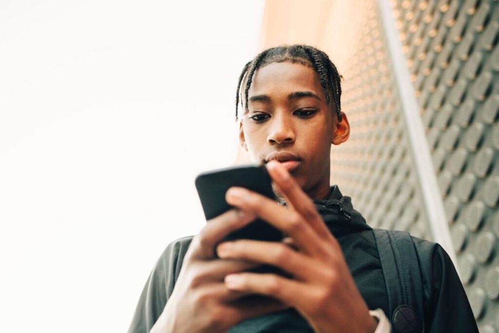 A young man holds a smartphone outside. He is looking down at the phone, engrossed.