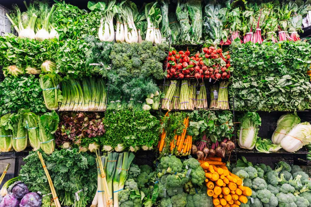 Fruits and vegetables are on display in a grocery store, including fennel, greens, onions, carrots, broccoli, and more.