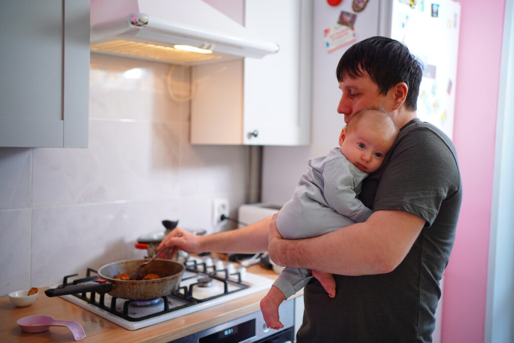 Parent cooks food at a gas stove while holding a baby.
