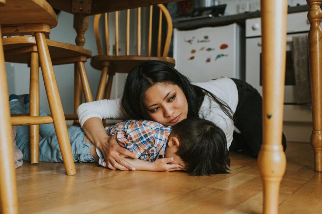 A mother holds and comforts a young boy who is crying face-down under a kitchen table.