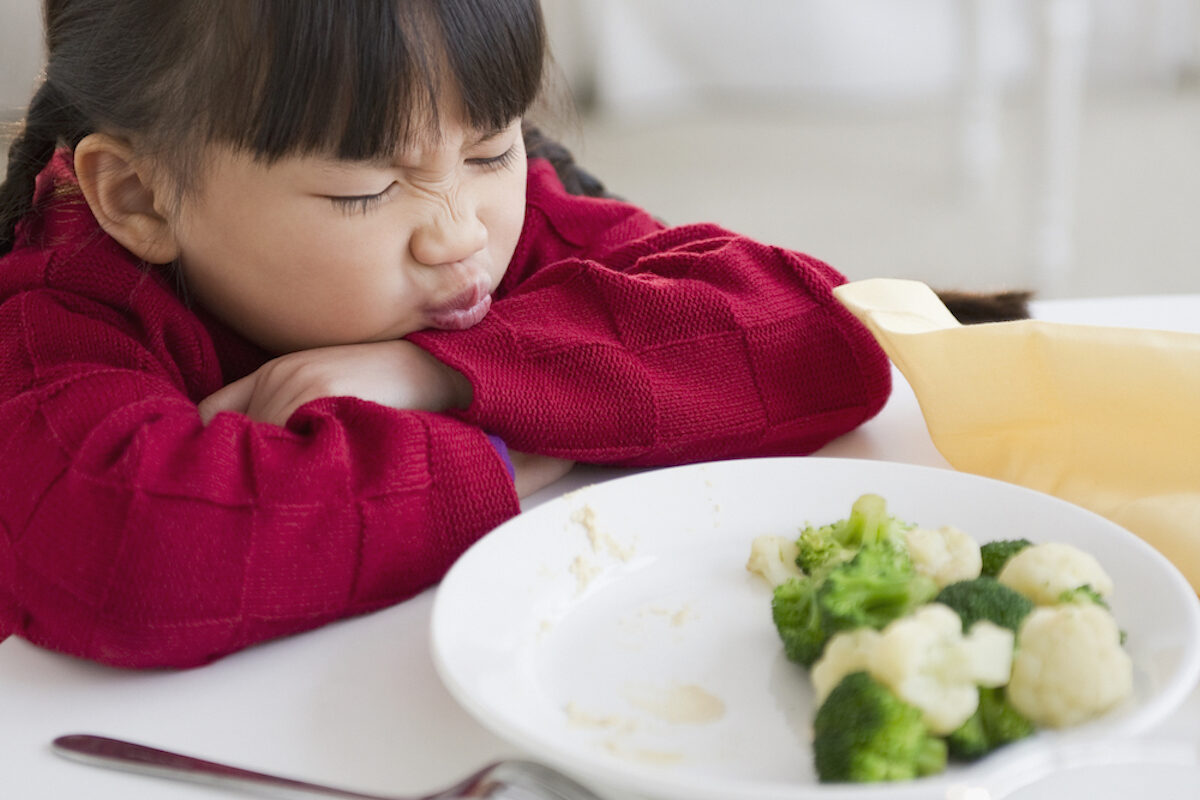 A child with their head resting on their arms makes a face at a bowl of broccoli and cauliflower.