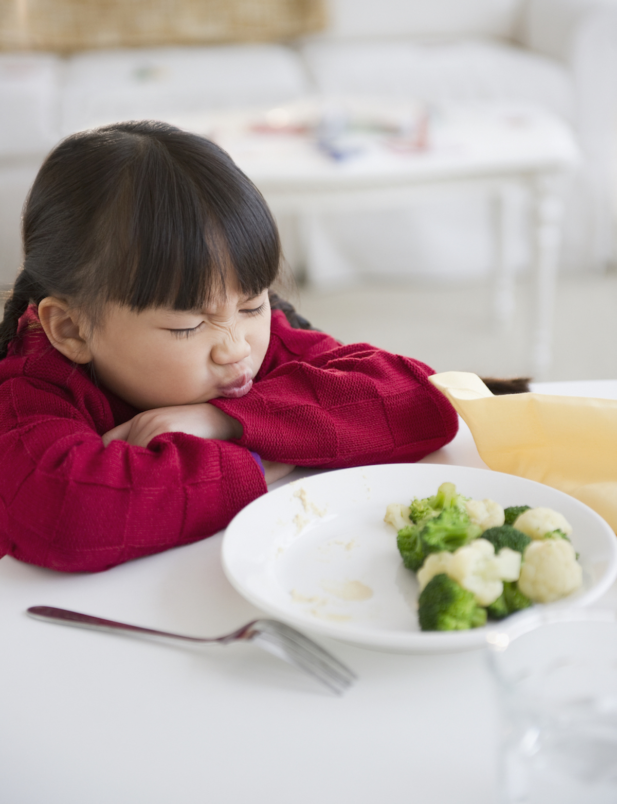 A child with their head resting on their arms makes a face at a bowl of broccoli and cauliflower.