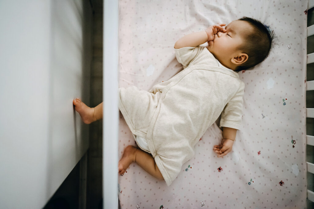 A baby sucks their thumb while sleeping on their back in a crib.