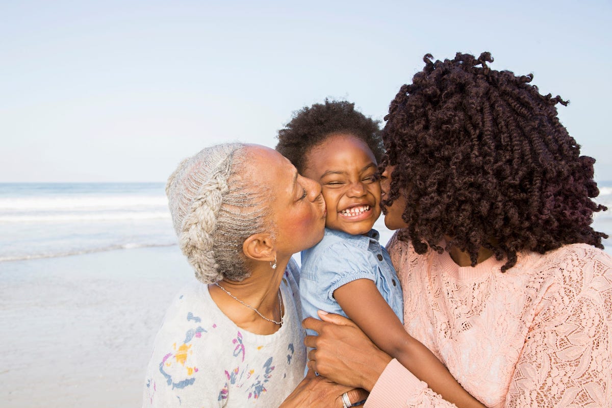 A Black mother and grandmother kiss both cheeks of a young child on a beach.