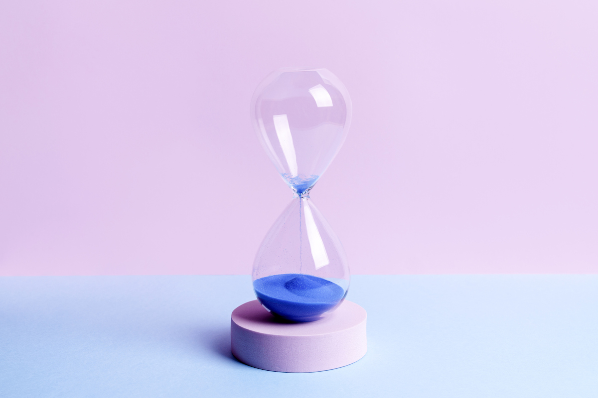 An hourglass filled with blue sand sits on a pink pedestal on a blue table. Most of the sand has fallen to the bottom chamber of the hourglass.