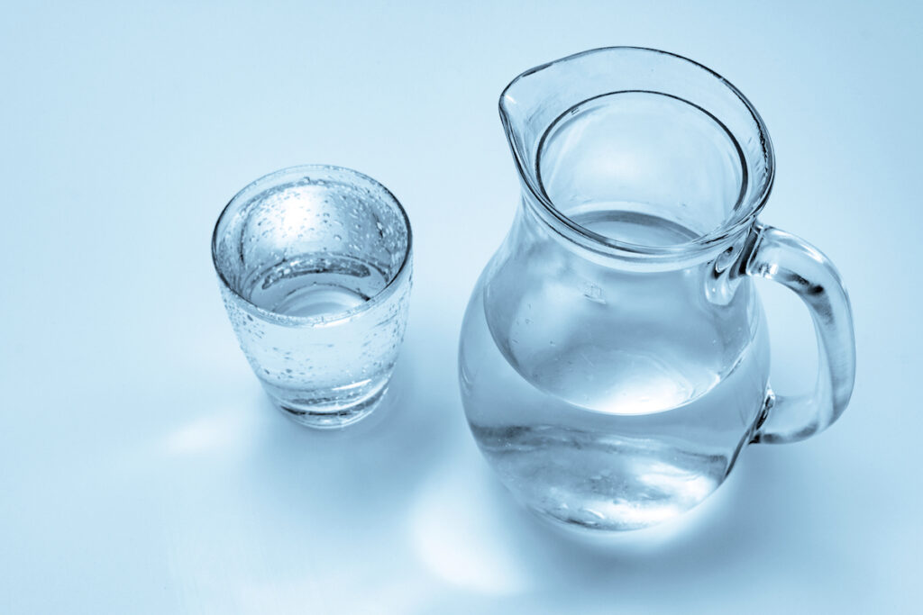 A glass jug of water and glass drinking glass are seen from overhead on a light blue background.