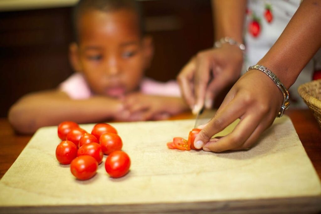 A parent chops tomatoes on a cutting board as a child looks on.