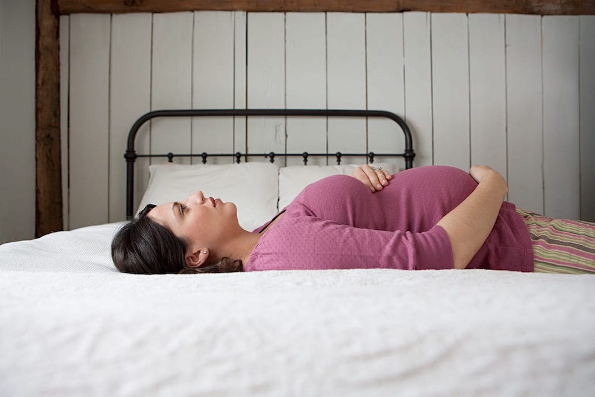 A pregnant person wearing a pink shirt lies across a bed, staring at the ceiling.