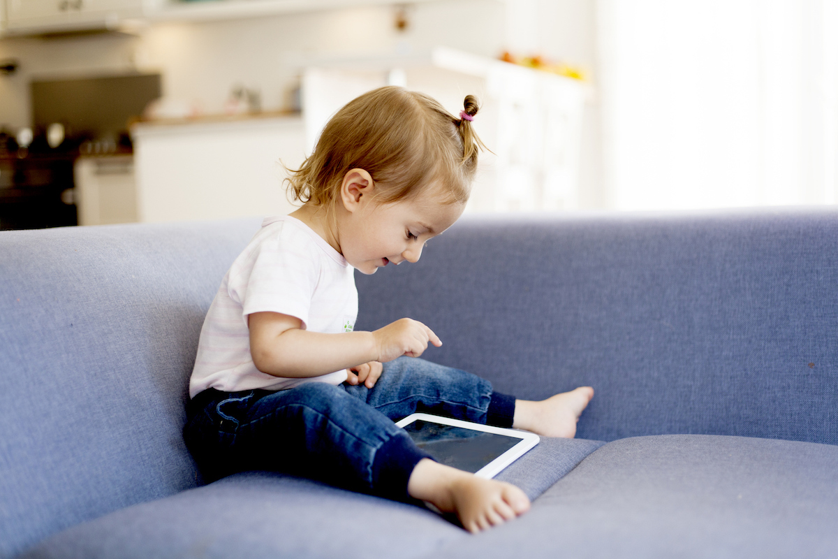 A toddler sits on a couch poking at an iPad and smiling.