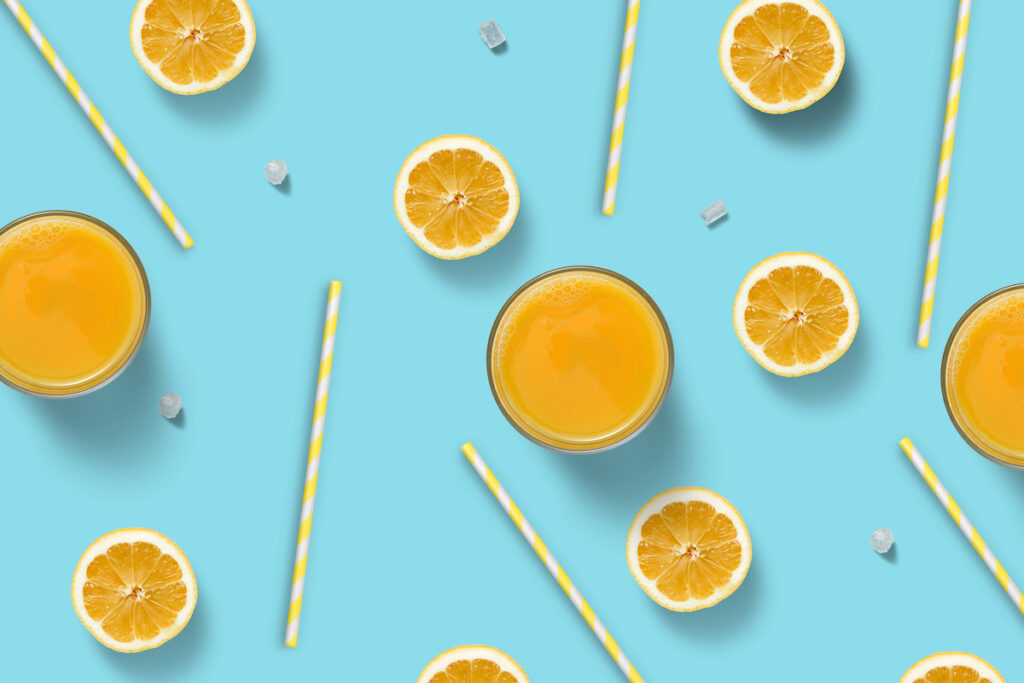 A bright blue background with an overhead arrangement of glasses of orange juice, orange straws, and orange slices.
