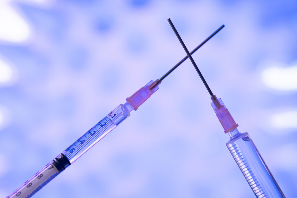 The needles of two vaccine syringes are crossed across a purple background.