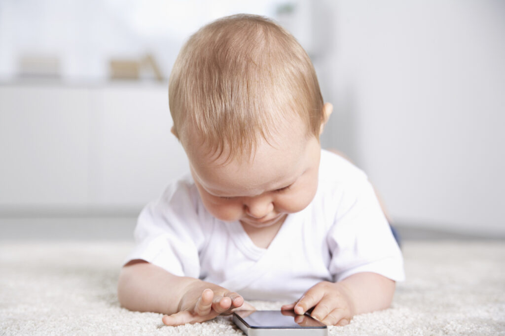 A small baby plays with a phone during tummy time and looks concerned.